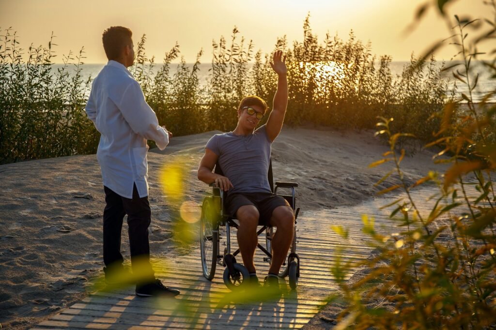 A young man using a wheelchair waving on a beach boardwalk while a support worker stands nearby