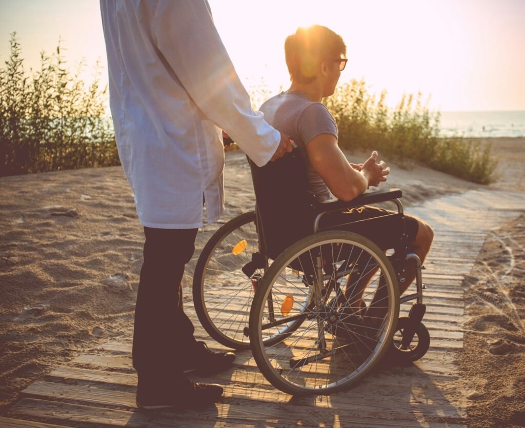 A man using a wheelchair on a coastal boardwalk at sunset with a support worker beside him